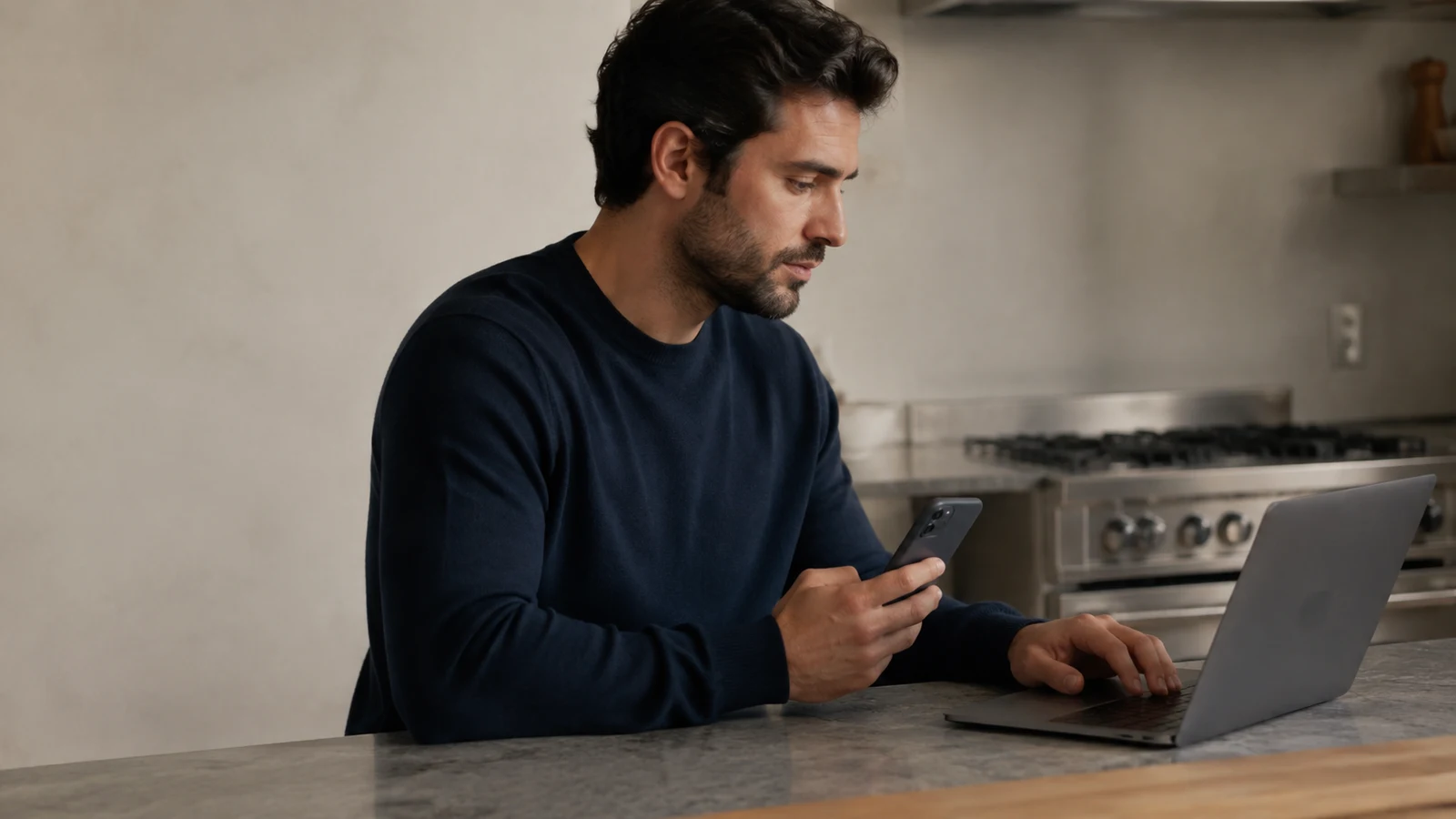 USDT shopping: A man holds a phone beside an open laptop on a stone kitchen counter.