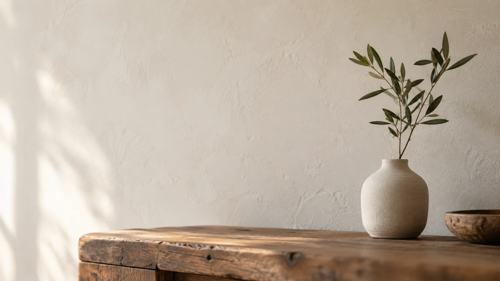 negative space photography: A vase with leafy branches sits on a wooden console beside a broad empty plaster wall.